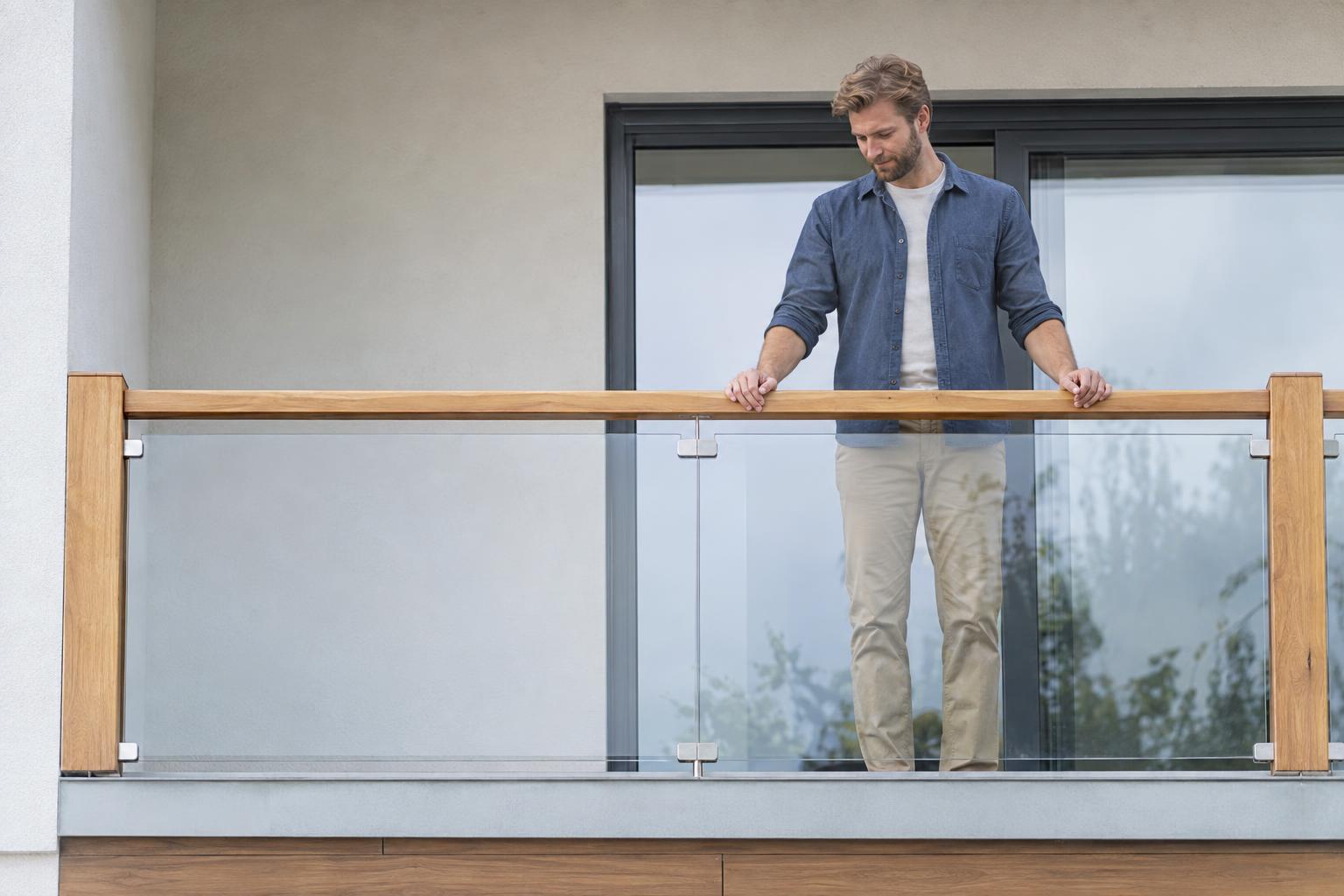 Un homme debout sur un balcon avec un garde-corps en verre et des mains sur la rambarde.